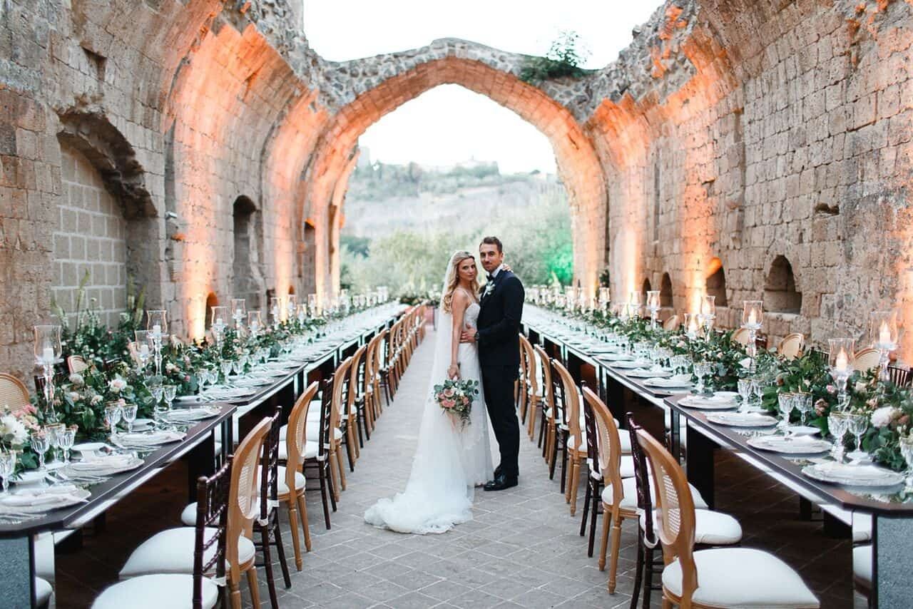 A couple between long candlelit tables under stone arches